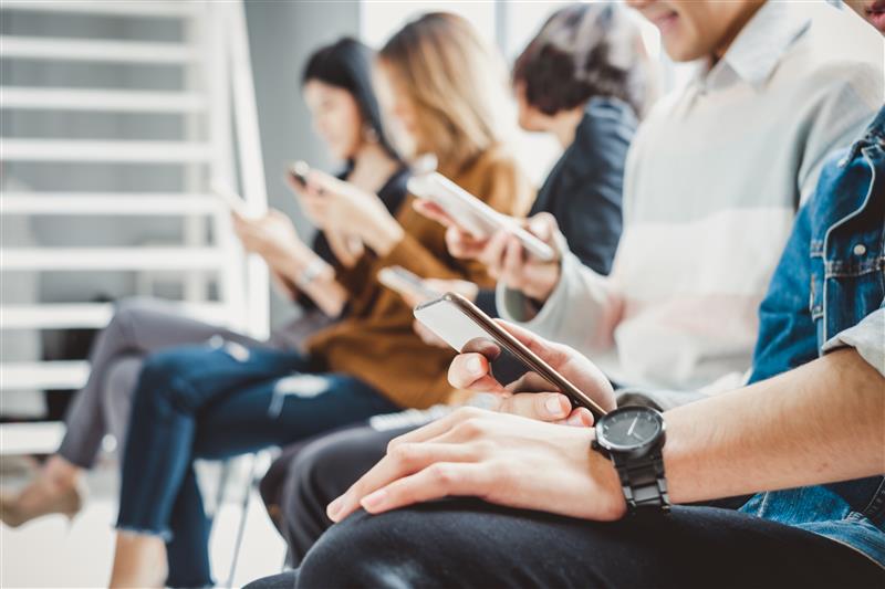 Group of young adults sitting in a row indoors, each focused on using their smartphones, highlighting modern digital habits and mobile engagement