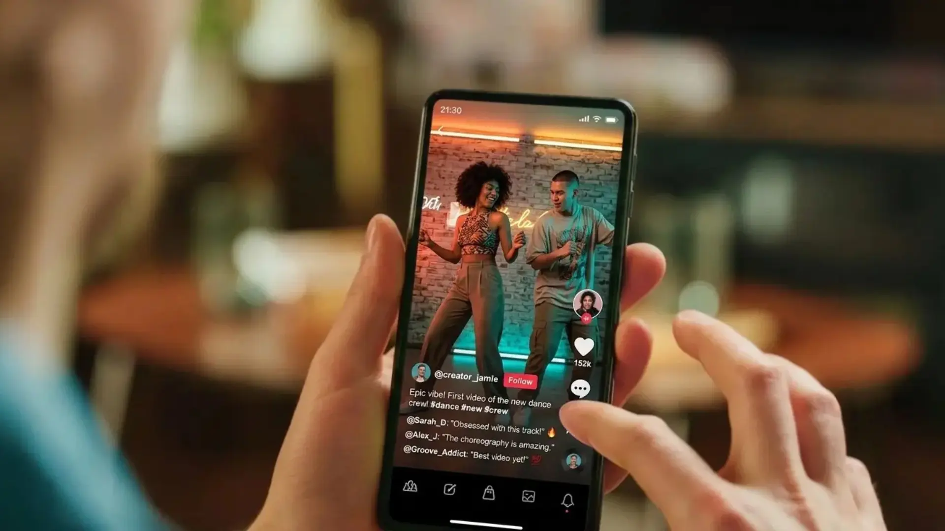 A close-up, over-the-shoulder shot of a person holding a smartphone in a dimly lit indoor setting. The phone screen displays a vertical social media video of two people—a man and a woman—dancing energetically in a studio with neon blue and orange lighting. The app interface is visible, featuring a "Follow" button,